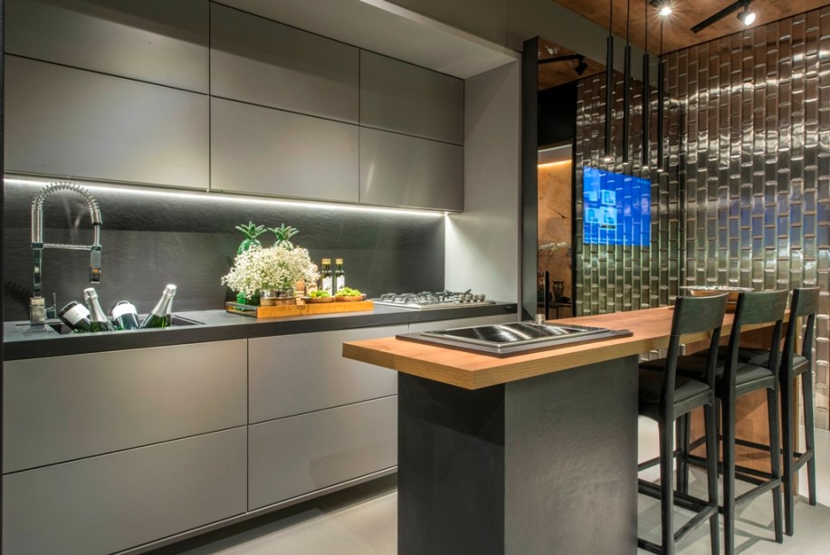 Cozinha Conceito - Ana Míriam Machado e Janice Diógenes. A dupla apostou no Dekton para modernizar o piso e as paredes. A esta base, agregaram o mobiliário cinza com portas de vidro prata. Além de automatizadas, as portas camuflam a geladeira, os fornos e as louças. A bancada recebe um tampo em Carvalho, mesma madeira que aquece o teto.