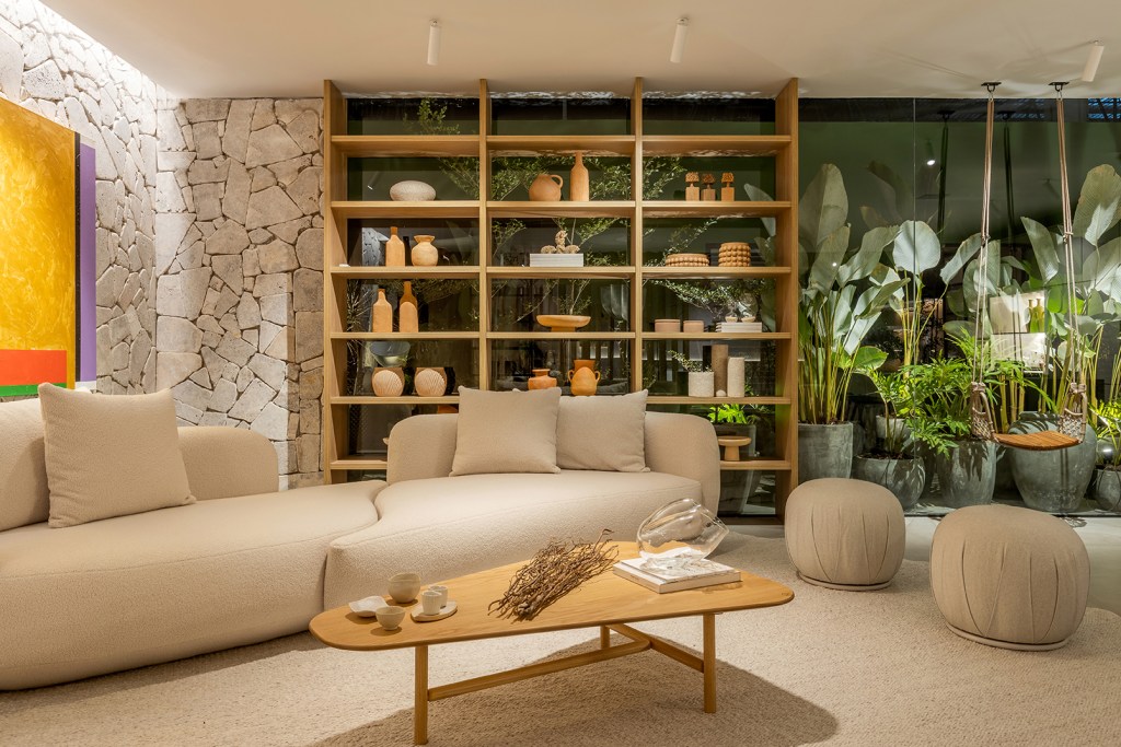 Cristiane Moussa - Refúgio Cenário. Projeto da CASACOR Goiás 2023. Na foto, sala de estar com plantas, sofá curvo e parede de pedra.