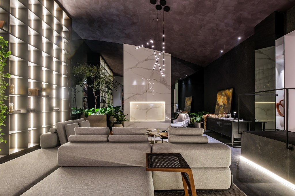 Kelly Leite - Living Principal Estar. Projeto da CASACOR Bolívia 2023. Na foto, sala de estar com lareira, estante iluminada, sofá e mesas.