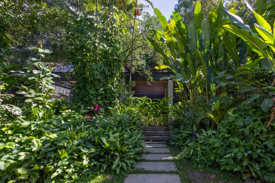 Catê Poli e Luis Porto - Jardim da Deca. Um jardim tropical, que mantém as espécies de Mata Atlântica já presentes na casa, e traz ainda mais folhagens verdes de plantas nativas, mas com um certo ar praiano, uma vez que incorpora a areia e as peças da Deca em exibição ali. Um espaço orgânico e natural, e, assim como todo o Pavilhão Deca, dedicado ao auto-cuidado, perfeito para relaxar e passar algumas horas apreciando a natureza. Móveis do designer Carlos Motta completam o ambiente criado pelos paisagistas Catê Poli e Luis Porto.