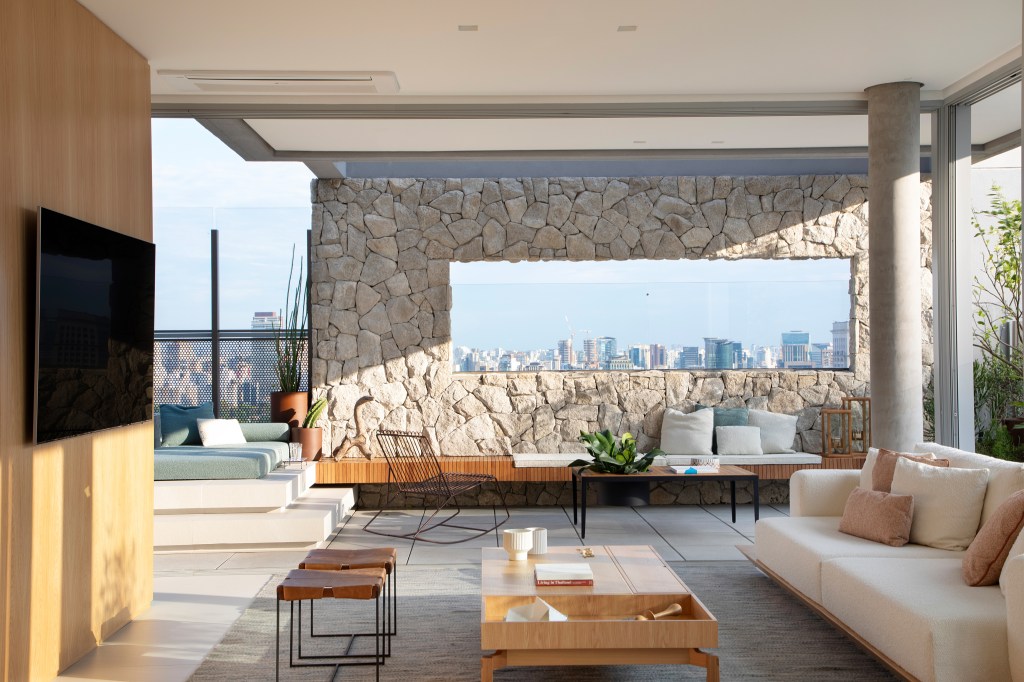 Skyline de São Paulo é emoldurado em apê assinado por Sabugosa Arquitetura. Na foto, Sala de estar integrada à varanda com muro de pedra e vista de SP.